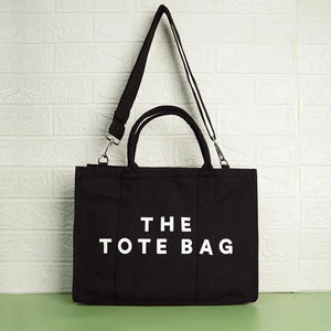 A black canvas tote bag with the text 'THE TOTE BAG' printed in white on the front side, featuring a shoulder strap and a zipper closure.