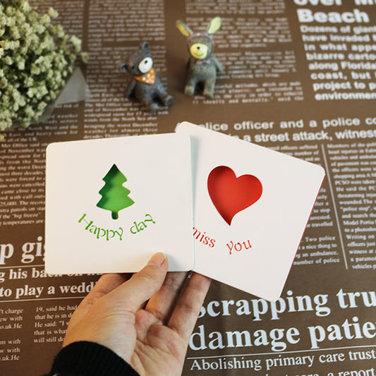 Two small greeting cards with a tree and heart design held by a hand on a newspaper background.