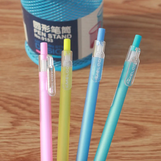 Four colorful pens with clear caps on a wooden surface, with a pen stand in the background.