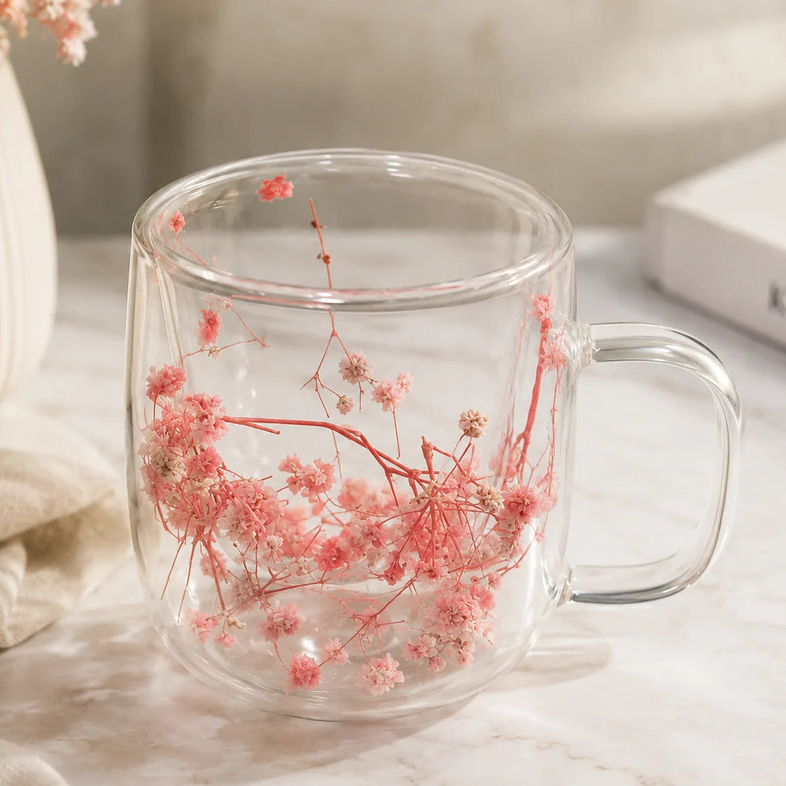 A clear glass mug with a handle, filled with dried pink flowers and red stems, set on a light marble surface with a neutral background and decorative props.