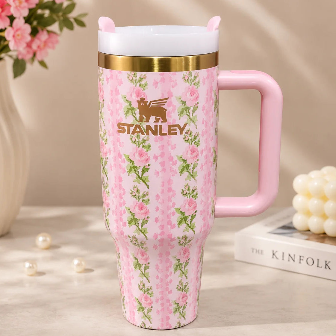 A pink Stanley insulated tumbler with a floral pattern, gold band, and white lid, styled on a tabletop with flowers, pearls, and a candle in the background.