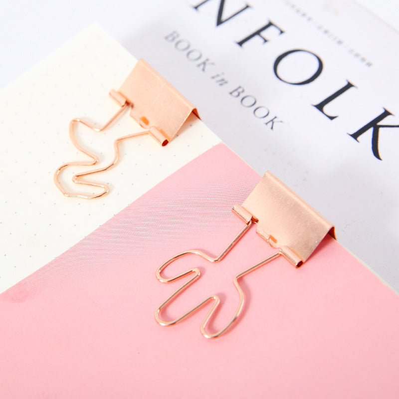Rose gold leaf-shaped paper clips on a pink surface with a book in the background.