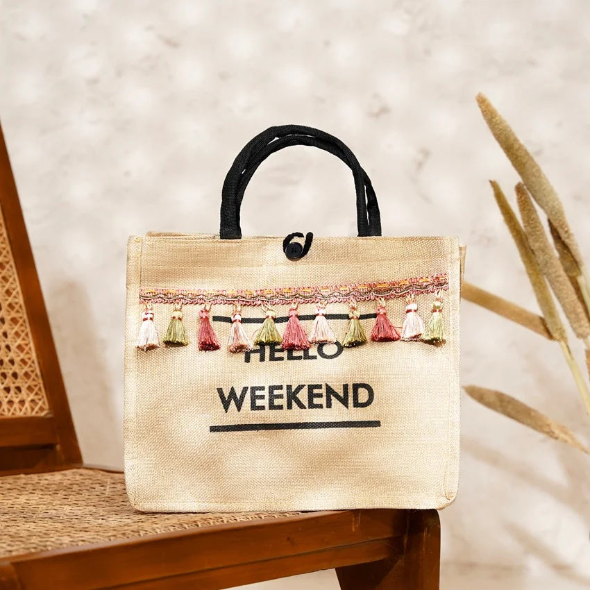 A beige tote bag with 'Hello Weekend' text printed in black on the front, decorated with multicolored pompoms and a black handle.