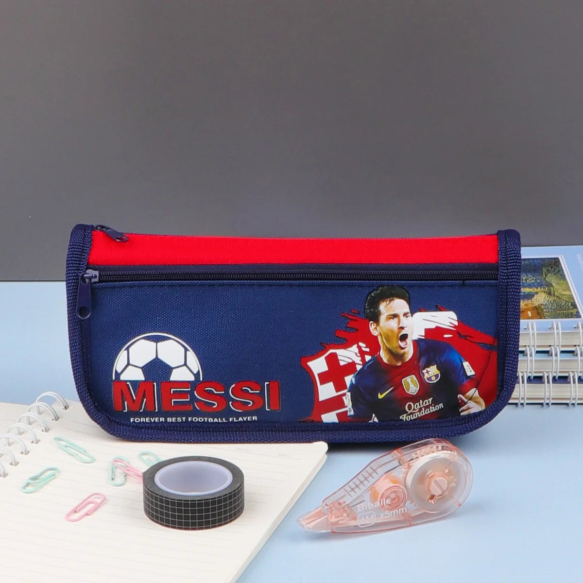 A pencil case with a zipper closure, featuring a soccer player on a blue background, and additional compartments. The pencil case is displayed in packaging labeled 'SCHOOL'.