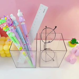 A transparent acrylic pen holder with a square shape, containing various colorful pens and a pair of glasses on a desk with decorative items around.