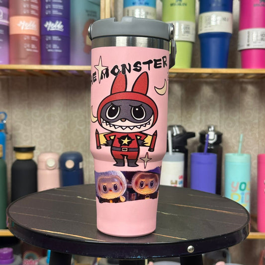 Pink tumbler with cartoon character design on a round black surface, with shelves of colorful tumblers in the background.