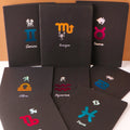 A collection of black notebooks with various star signs printed on the covers, including Aries, Taurus, Gemini, Scorpio, Libra, and Pisces. Each notebook has a black spine and a different colored cover for each sign.