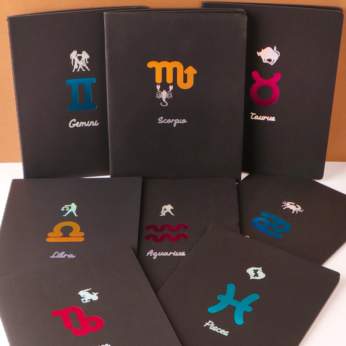 A collection of black notebooks with various star signs printed on the covers, including Aries, Taurus, Gemini, Scorpio, Libra, and Pisces. Each notebook has a black spine and a different colored cover for each sign.