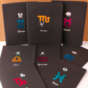 A collection of black notebooks with various star signs printed on the covers, including Aries, Taurus, Gemini, Scorpio, Libra, and Pisces. Each notebook has a black spine and a different colored cover for each sign.