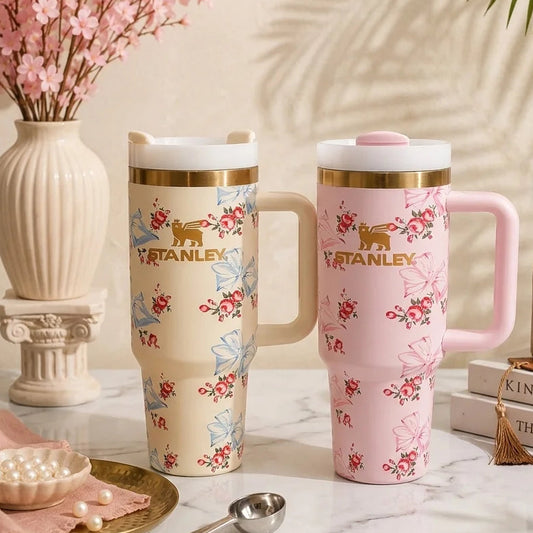 Two Stanley travel mugs with lids, one cream and one pink, decorated with floral and butterfly patterns, displayed on a marble surface with decorative props.