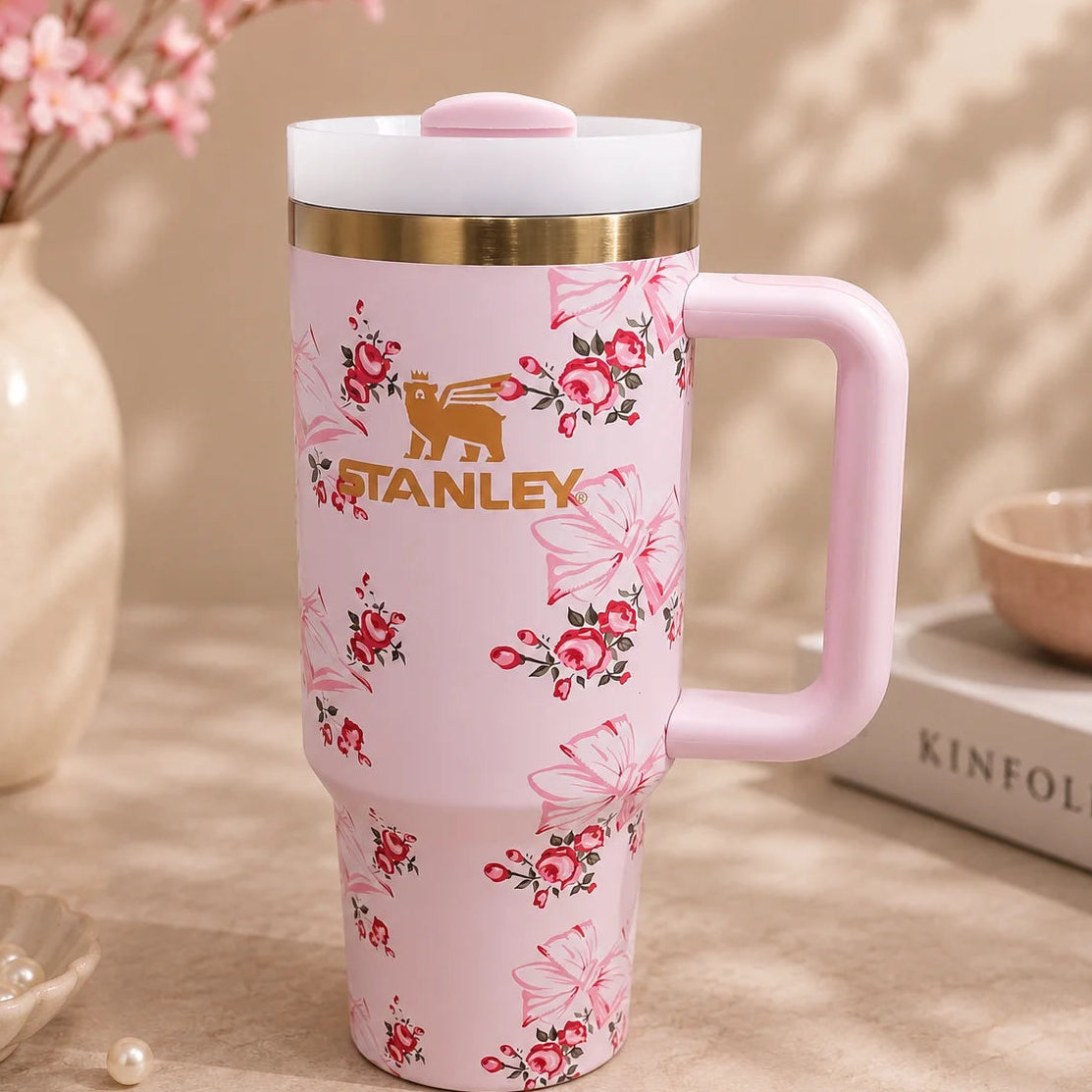 A pink Stanley travel tumbler with a floral and bow pattern, featuring the gold Stanley bear logo and text on the front. The tumbler has a light pink handle, a white lid with a pink top, and is set on a beige surface with decorative props including a vase of pink flowers, pearls, and a book.
