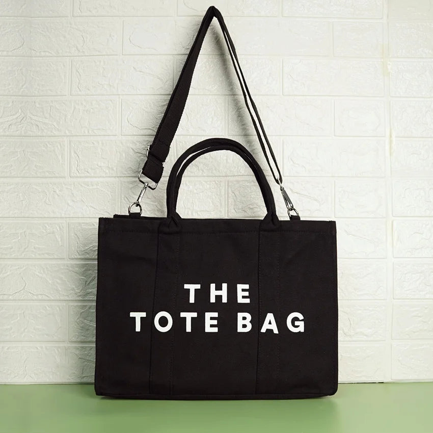 A black canvas tote bag with the text 'THE TOTE BAG' printed in white on the front side, featuring a shoulder strap and a zipper closure.