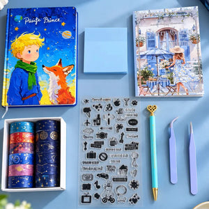 A flat-lay arrangement of stationery and craft items on a light blue surface: a blue 'Le Petit Prince' metal tin, a printed art card, a small blue notebook, washi tape rolls in a white box, a sheet of clear stamps, a turquoise pen with a gold heart, and two lavender tweezers.