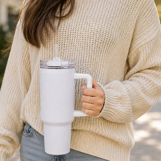 A person wearing a cream knit sweater and light blue jeans holds a white insulated travel mug with a clear lid outdoors on a paved walkway.