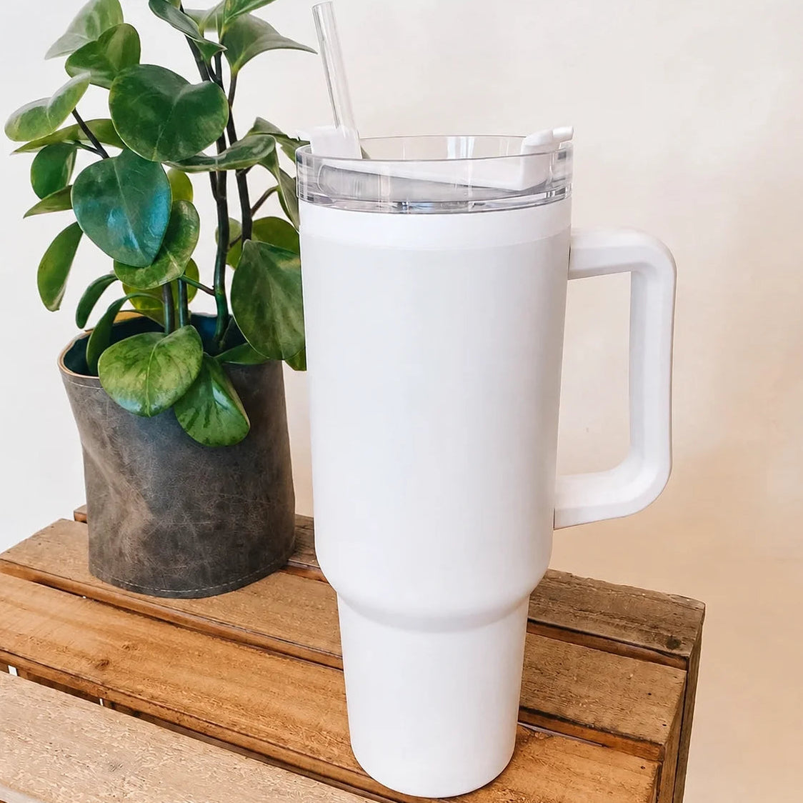 A tall white insulated tumbler with a clear lid and straw sits on a wooden crate next to a potted green plant against a plain light beige wall.