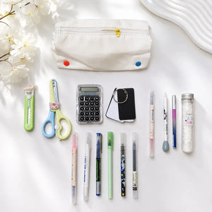 A styled flat-lay of school and office supplies on a white surface, including a white fabric pencil pouch with colorful snaps, scissors, a utility knife, a calculator, a small black card holder, multiple pens, and a clear bottle.