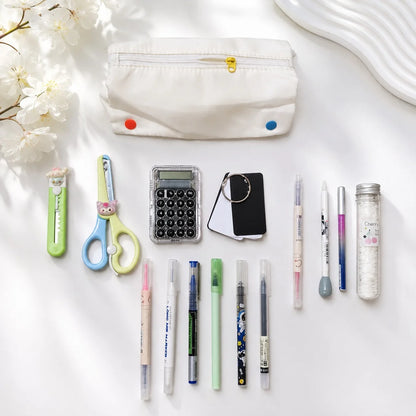 A styled flat-lay of school and office supplies on a white surface, including a white fabric pencil pouch with colorful snaps, scissors, a utility knife, a calculator, a small black card holder, multiple pens, and a clear bottle.