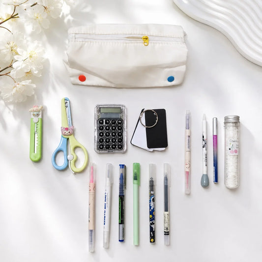 A styled flat-lay of school and office supplies on a white surface, including a white fabric pencil pouch with colorful snaps, scissors, a utility knife, a calculator, a small black card holder, multiple pens, and a clear bottle.