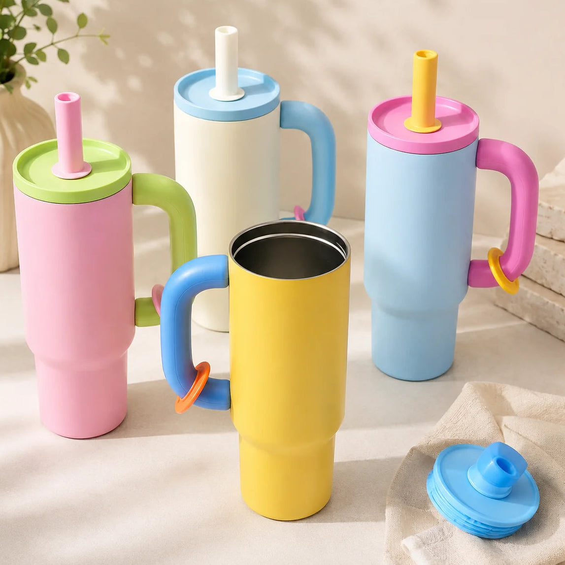 Four handled travel tumblers with straw lids in pastel pink, cream, light blue, and yellow, arranged on a light beige surface with soft shadows. One blue lid is placed separately in the lower right corner.