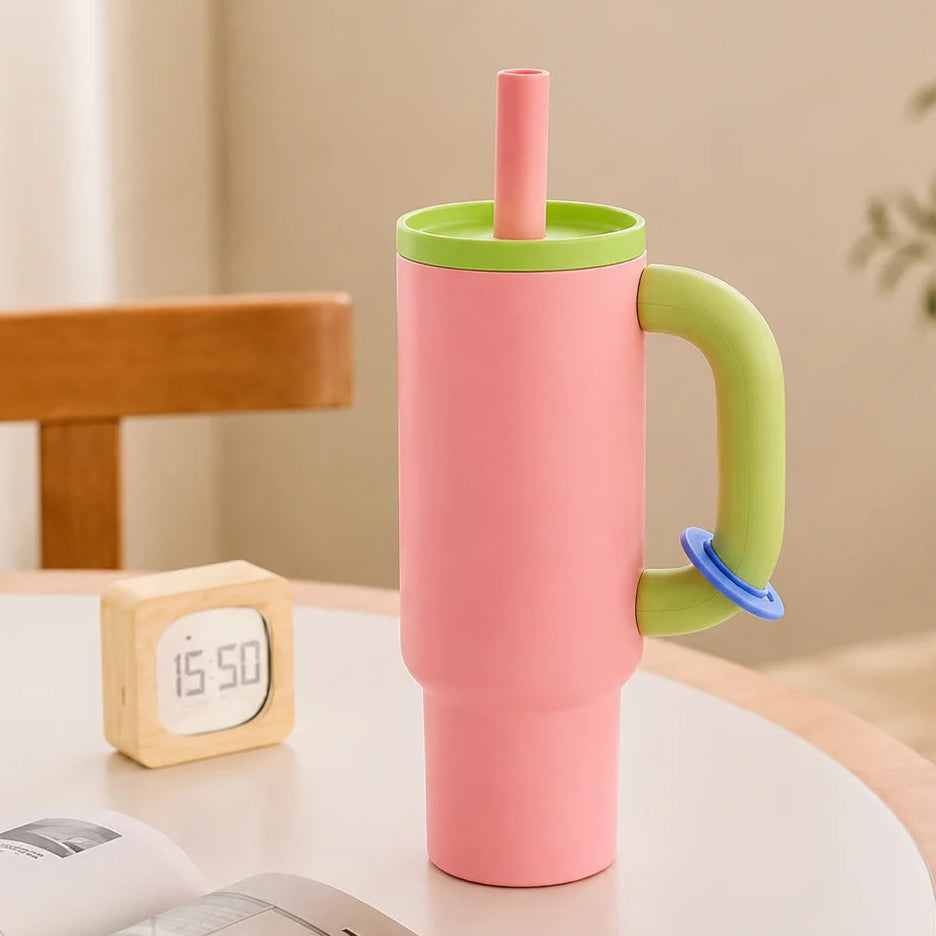 A pink handled tumbler with a light green lid and handle, plus a pink straw and small blue accent, standing on a round table in a softly lit room. A small wooden clock, open book, and chair are visible around it.