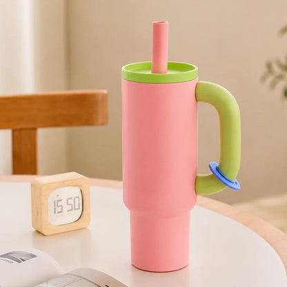 A pink handled tumbler with a light green lid and handle, plus a pink straw and small blue accent, standing on a round table in a softly lit room. A small wooden clock, open book, and chair are visible around it.