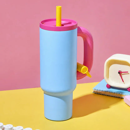 A pastel blue insulated travel tumbler with a pink lid and handle, yellow straw, and small yellow side spout, styled on a yellow desk with a pink background, keyboard, mouse, notebook, and retro clock.