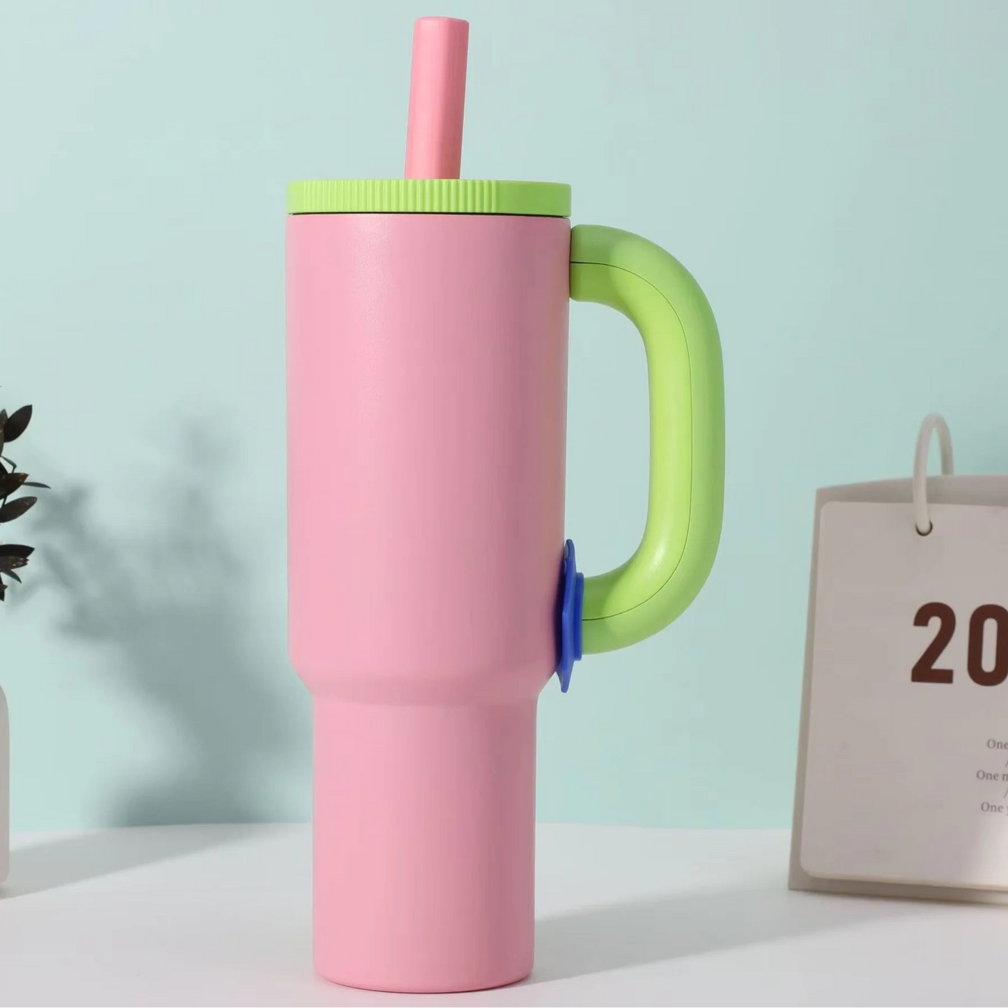 A pastel pink insulated tumbler with a bright green handle, matching green lid, and pink straw, shown on a white tabletop against a pale blue wall. A small vase with leaves and a partial desk calendar are visible as styled props.
