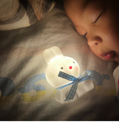 Child sleeping next to a white bear-shaped night light with a blue ribbon on a textured surface.