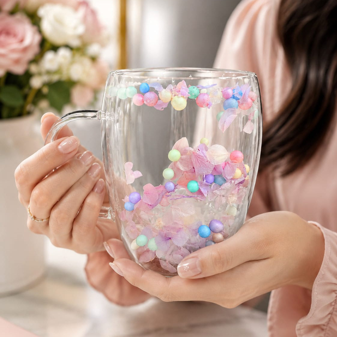 Clear mug with colorful beads held by a person, with flowers in the background