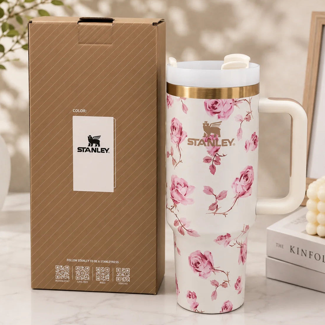 A Stanley travel mug with a pink rose floral pattern on a white background, shown next to its brown branded box on a marble surface with decorative props.