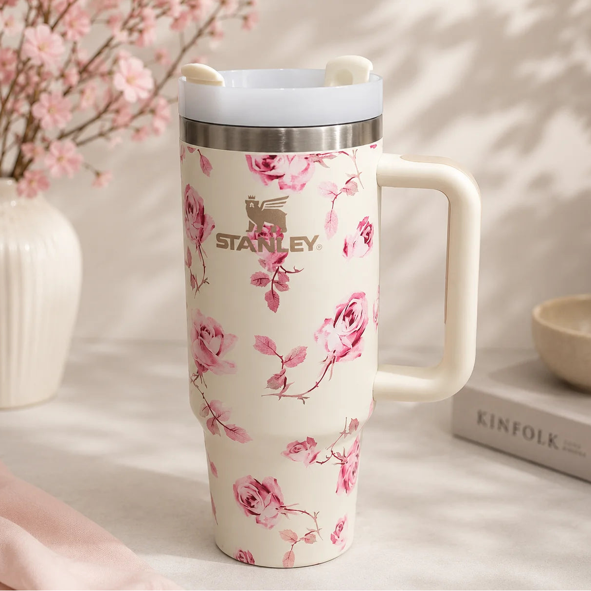 A Stanley travel mug with a pink floral print on a cream background, featuring a stainless steel rim, a cream handle, and a matching lid with small ear-like details. The mug is styled on a light surface with soft shadows, a vase of pink blossoms, and a book in the background.