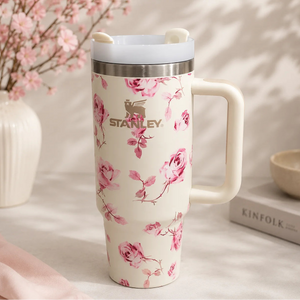 A Stanley travel mug with a pink floral print on a cream background, featuring a stainless steel rim, a cream handle, and a matching lid with small ear-like details. The mug is styled on a light surface with soft shadows, a vase of pink blossoms, and a book in the background.