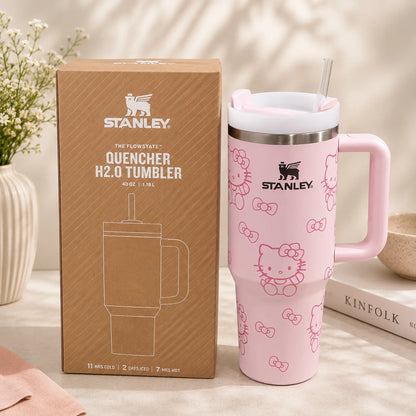 A pink Stanley Hello Kitty insulated tumbler with a handle and straw stands beside its brown branded retail box on a light tabletop. The tumbler has a white lid, a stainless steel rim, and pink Hello Kitty line-art patterns.