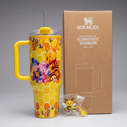 A Stanley Flowstate tumbler with a Winnie the Pooh honeycomb pattern and character illustrations, shown next to its brown cardboard packaging box and a small bee accessory on a gray background.