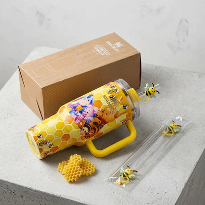 A yellow honeybee-themed insulated tumbler with a yellow handle, shown with its brown branded box, a small honeycomb piece, and two bee accessories on a light stone surface.