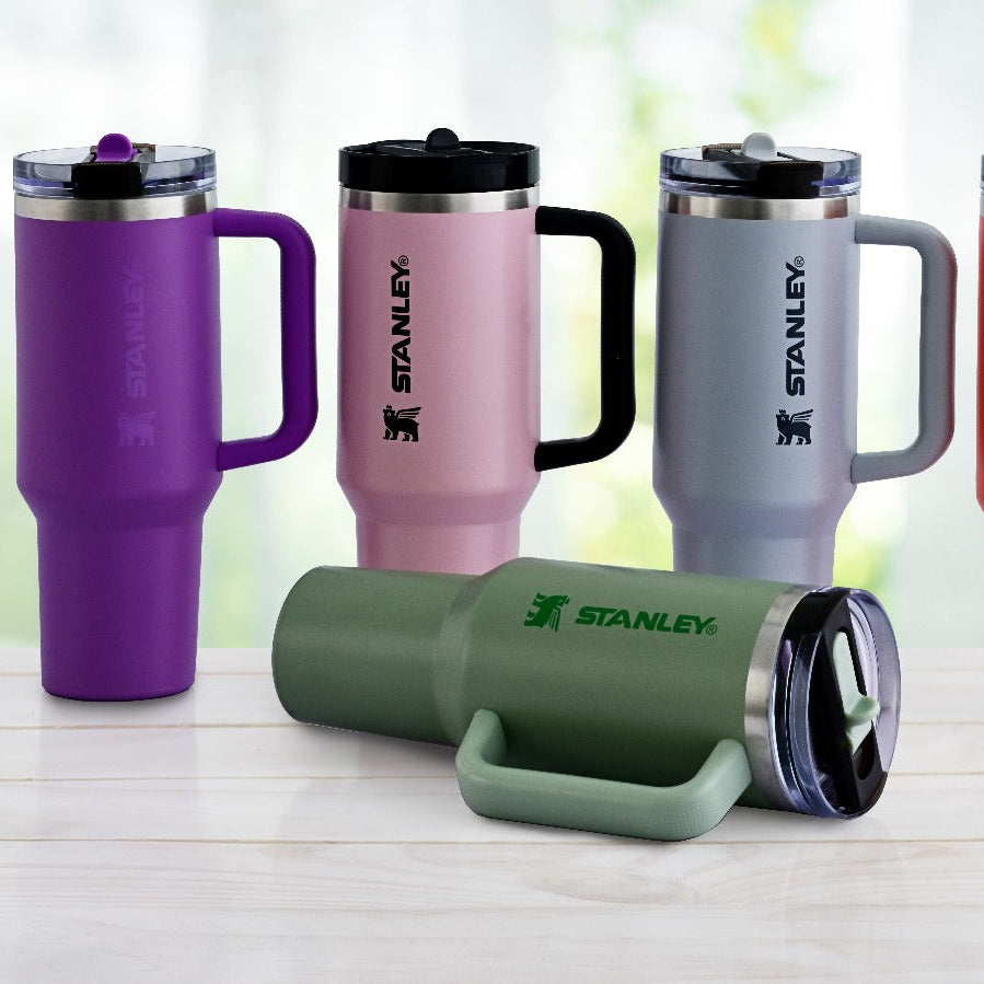 Five Stanley travel mugs in different colors on a white surface with a blurred green and brown background.
