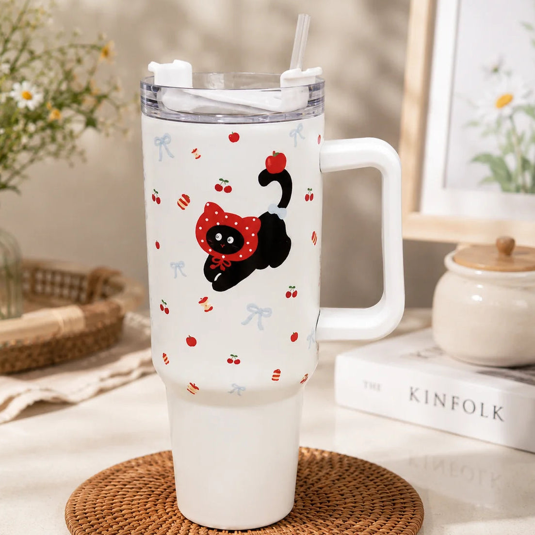 A white handled tumbler with a clear lid and straw, decorated with a black cat wearing a red polka-dot bow and small red cherry motifs, set on a woven coaster in a styled indoor scene.