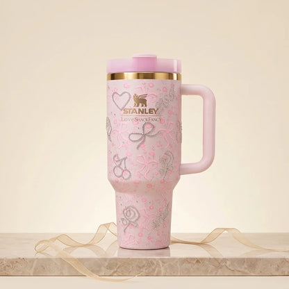 A pink Stanley travel tumbler with a matching lid, gold rim, and silver glitter patterns, displayed on a marble surface with a beige ribbon.