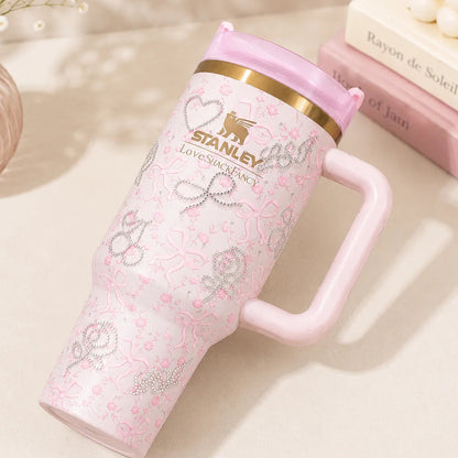A pink Stanley Love in Space handled tumbler with a gold band and pink lid, styled on a beige surface next to books and a vase.
