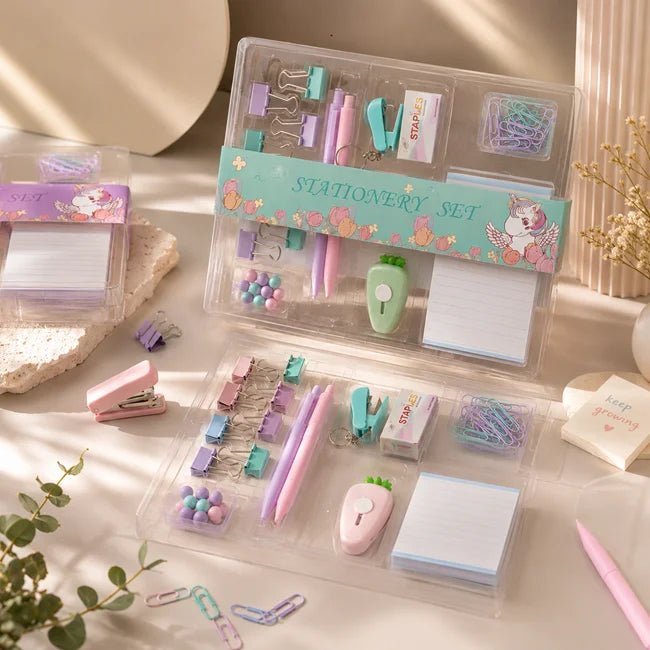 A pastel stationery set displayed in clear plastic organizer cases on a light tabletop. The set includes a pink stapler, purple and pink pens, clips, paper clips, sticky notes, a small apple-shaped eraser, and other stationery accessories.