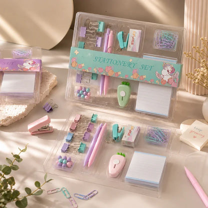 A pastel stationery set displayed in clear plastic organizer cases on a light tabletop. The set includes a pink stapler, purple and pink pens, clips, paper clips, sticky notes, a small apple-shaped eraser, and other stationery accessories.