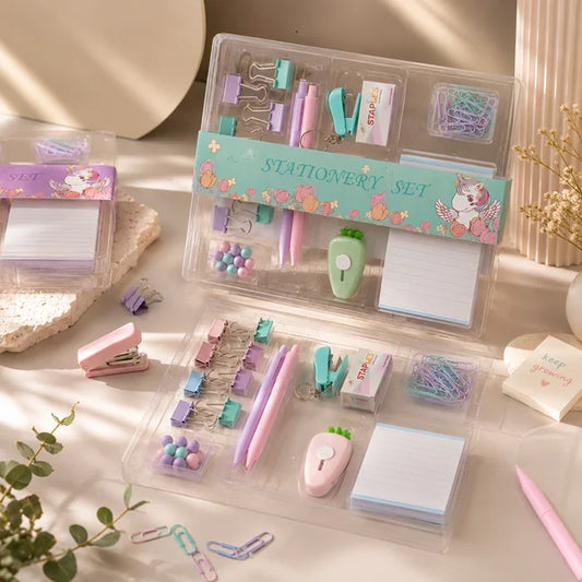 A pastel stationery set displayed in clear plastic organizer cases on a light tabletop. The set includes a pink stapler, purple and pink pens, clips, paper clips, sticky notes, a small apple-shaped eraser, and other stationery accessories.