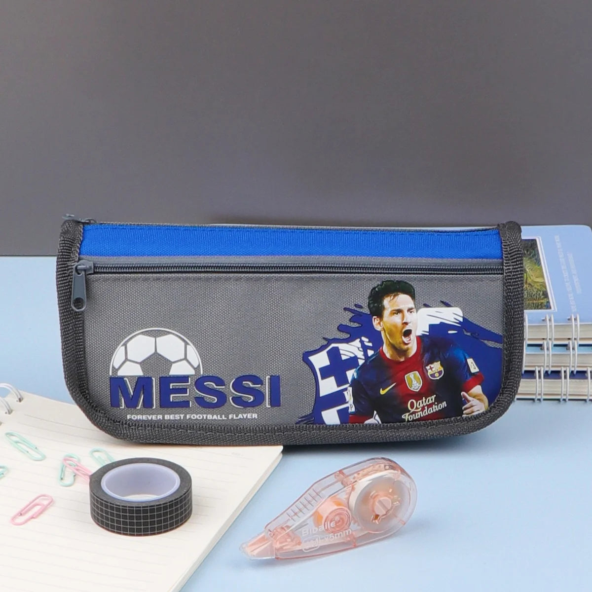A pencil case with a zipper closure, featuring a soccer player on a blue background, and additional compartments. The pencil case is displayed in packaging labeled 'SCHOOL'.
