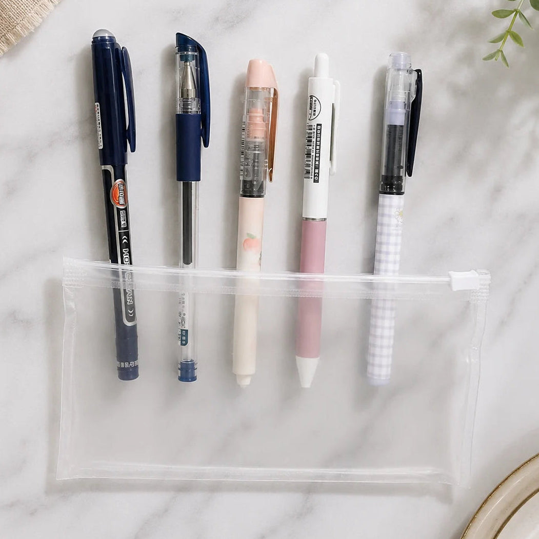 A clear zippered pencil pouch laid on a white marble surface with five pens arranged beside it. The scene includes a small green plant leaf in the top left and part of a beige plate in the top right.