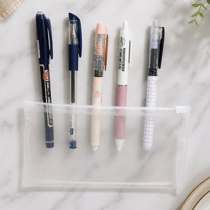 A clear zippered pencil pouch laid on a white marble surface with five pens arranged beside it. The scene includes a small green plant leaf in the top left and part of a beige plate in the top right.