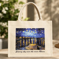 A cream canvas tote bag with double top handles, featuring a printed graphic of Van Gogh's 'Starry sky over the river Rhine' and the text 'Starry sky over the river Rhine' below it. The bag is displayed on a wooden table with a blurred background of greenery and warm sunlight.