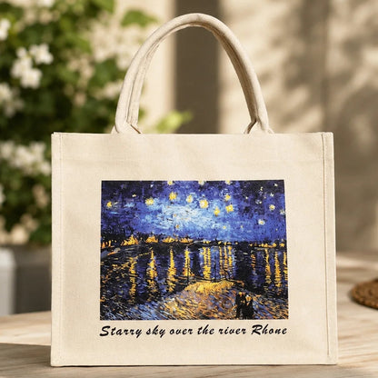 A cream canvas tote bag with double top handles, featuring a printed graphic of Van Gogh's 'Starry sky over the river Rhine' and the text 'Starry sky over the river Rhine' below it. The bag is displayed on a wooden table with a blurred background of greenery and warm sunlight.