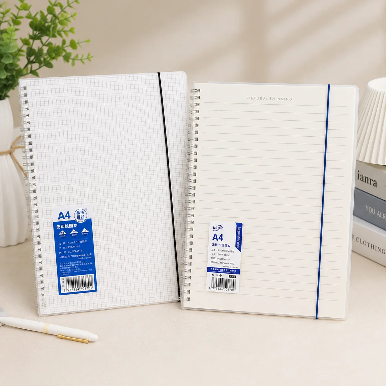 Two A4 notebooks on a light beige desk. One notebook shows a white grid pattern cover with a blue and white sticker, and the other shows a white lined interior with blue lines and a blue elastic band.