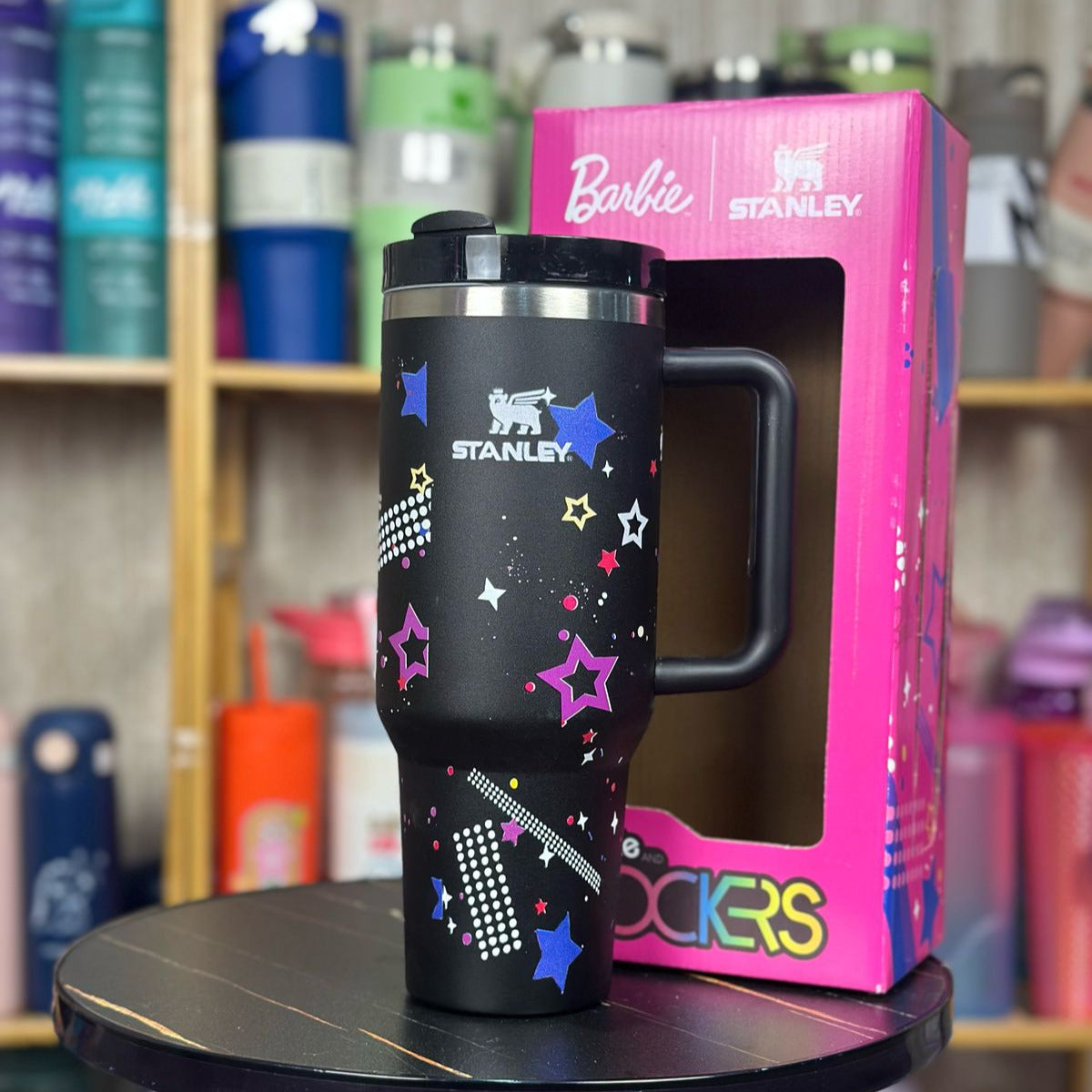 Black Stanley tumbler with colorful designs on a pink Barbie Stanley packaging, placed on a black stool with a blurred background of shelves with various items.
