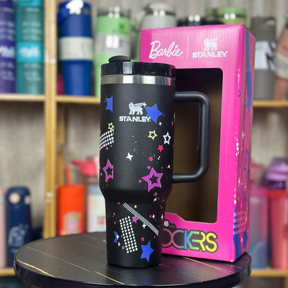Black Stanley tumbler with colorful designs on a pink Barbie Stanley packaging, placed on a black stool with a blurred background of shelves with various items.